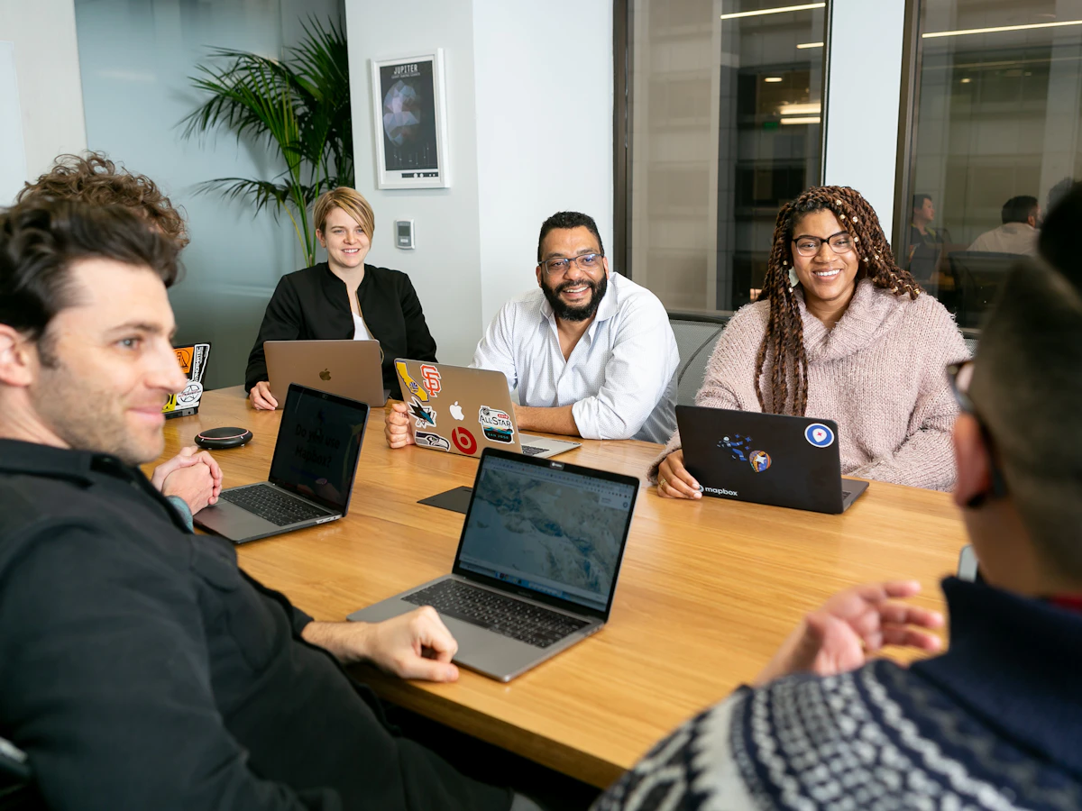 Cinq collaborateurs en réunion autour d'une table de bureau, ordinateurs portables ouverts.